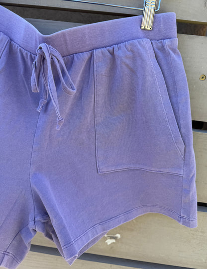 Lavender drawstring shorts with a side pocket, hanging from a wooden hanger on a weathered gray wooden slat surface.