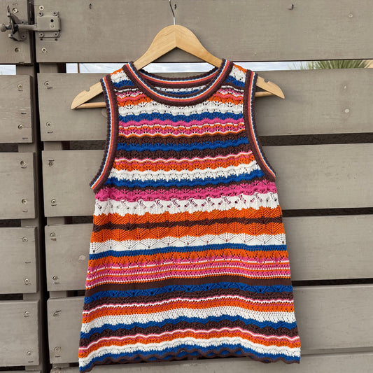 A sleeveless knit top with horizontal wavy stripes in orange, red, pink, blue, and white, hanging on a wooden hanger against a gray wooden fence under a slatted wooden roof.