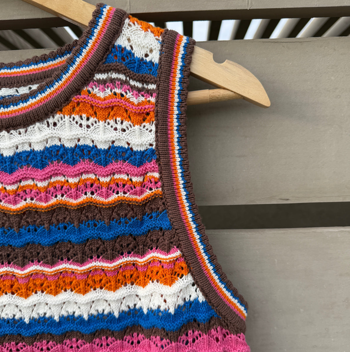 A sleeveless knit top with horizontal multicolor stripes in orange, brown, white, blue, pink, and red, hanging on a wooden hanger against a gray wooden slat background.