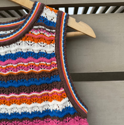 A sleeveless knit top with horizontal multicolor stripes in orange, brown, white, blue, pink, and red, hanging on a wooden hanger against a gray wooden slat background.