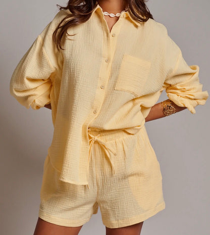 A woman wearing a pale yellow textured cotton romper with a tie waist and chest pocket, photographed against a plain light gray studio background.