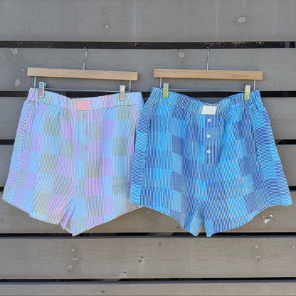 Two pairs of plaid shorts hanging on a wooden fence with a striped awning above.