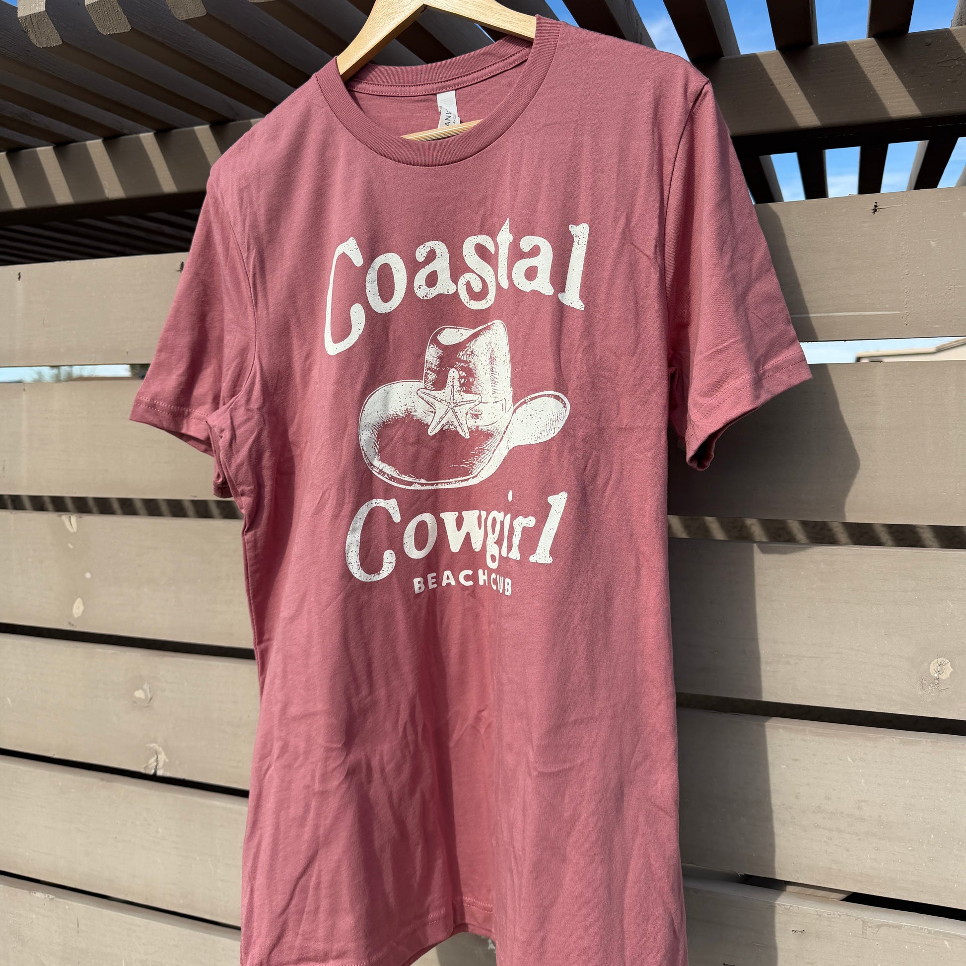 Mauve t-shirt with 'Coastal Cowgirl Ranch' print on a wooden surface