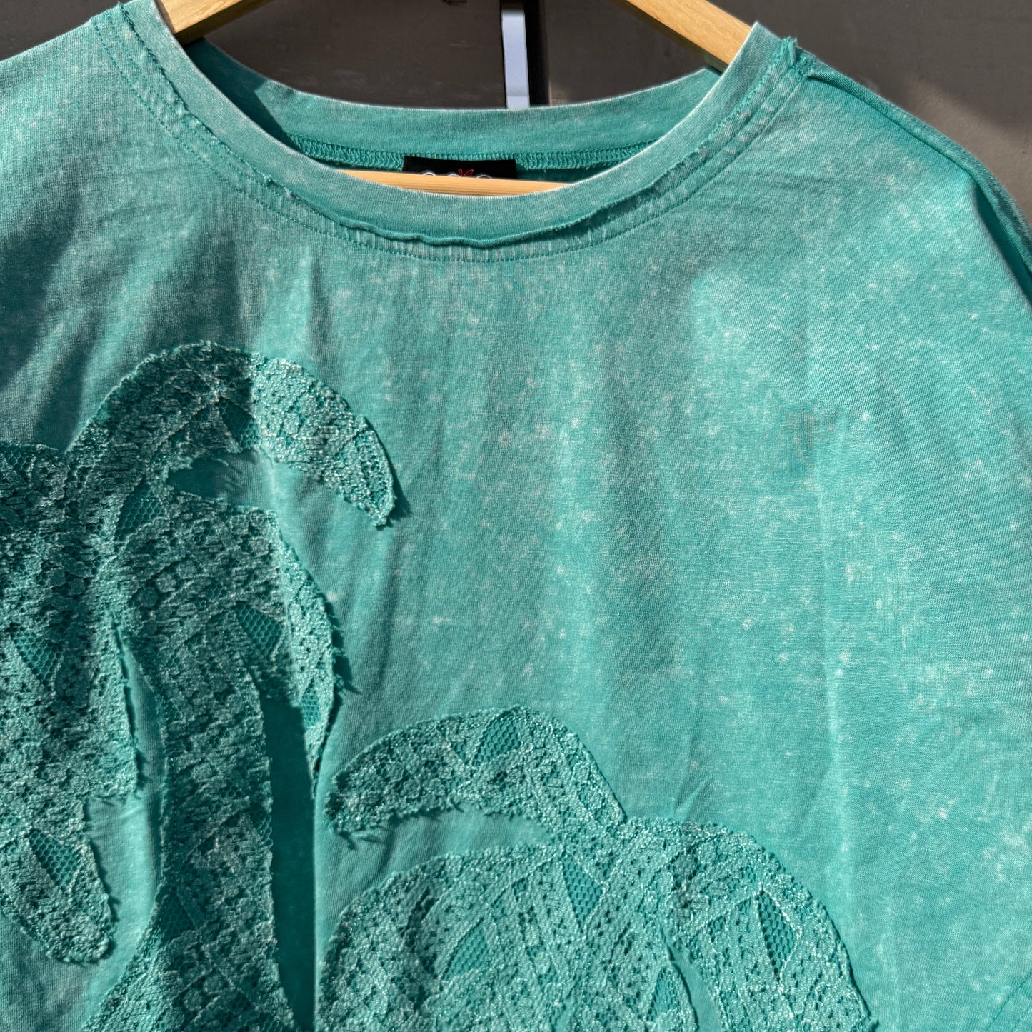 Teal t-shirt with lace detailing on a hanger against a dark background