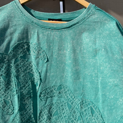 Teal t-shirt with lace detailing on a hanger against a dark background