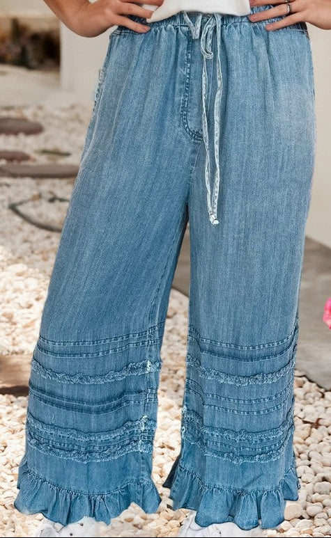 Blue denim wide-leg pants with ruffled bottom hem worn by a person on a pebbled surface.