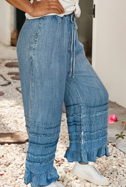 Denim wide-leg pants with ruffled bottom hem worn by a person on a pebbled surface.