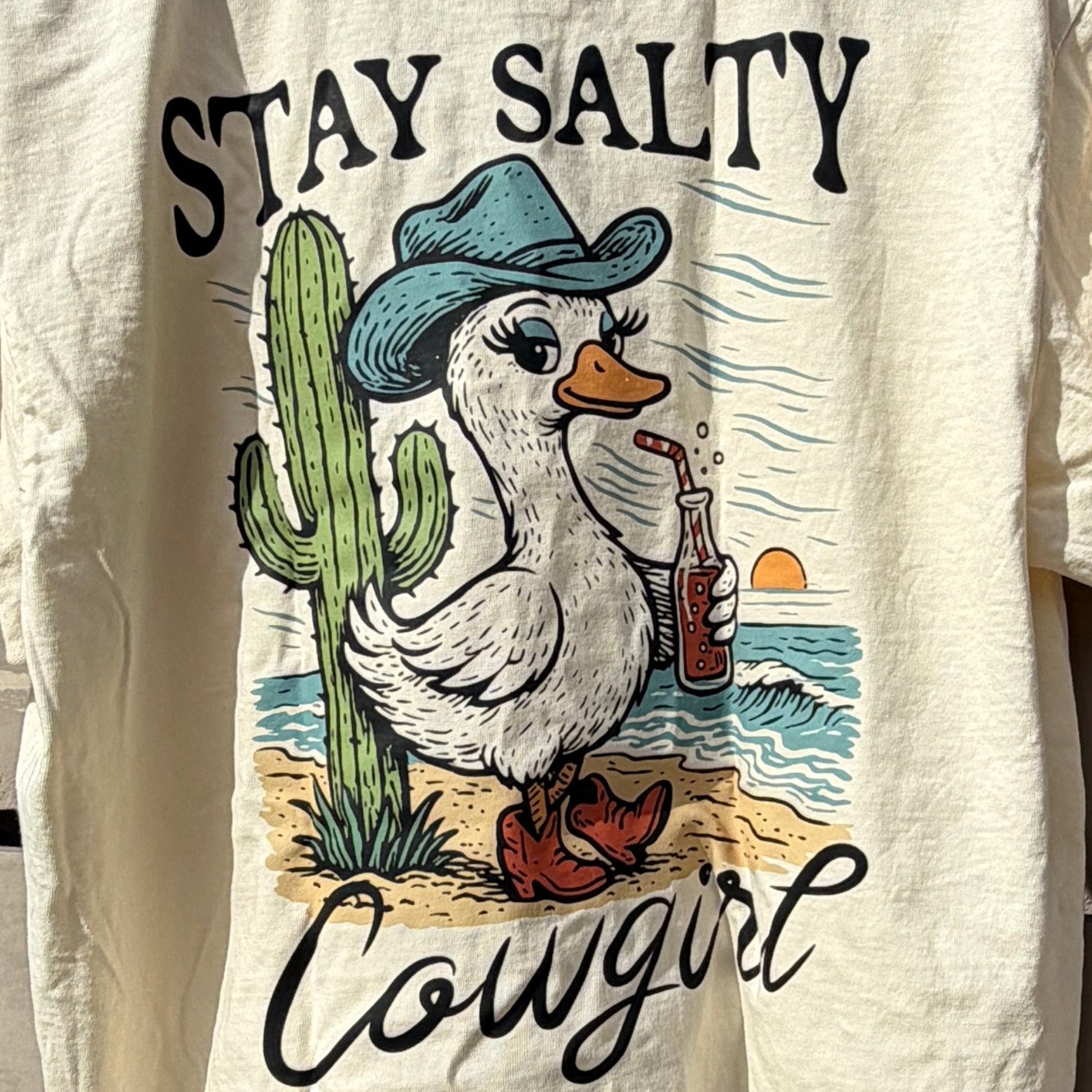 Beige tshirt with graphic design and text on a wooden deck with blue sky background